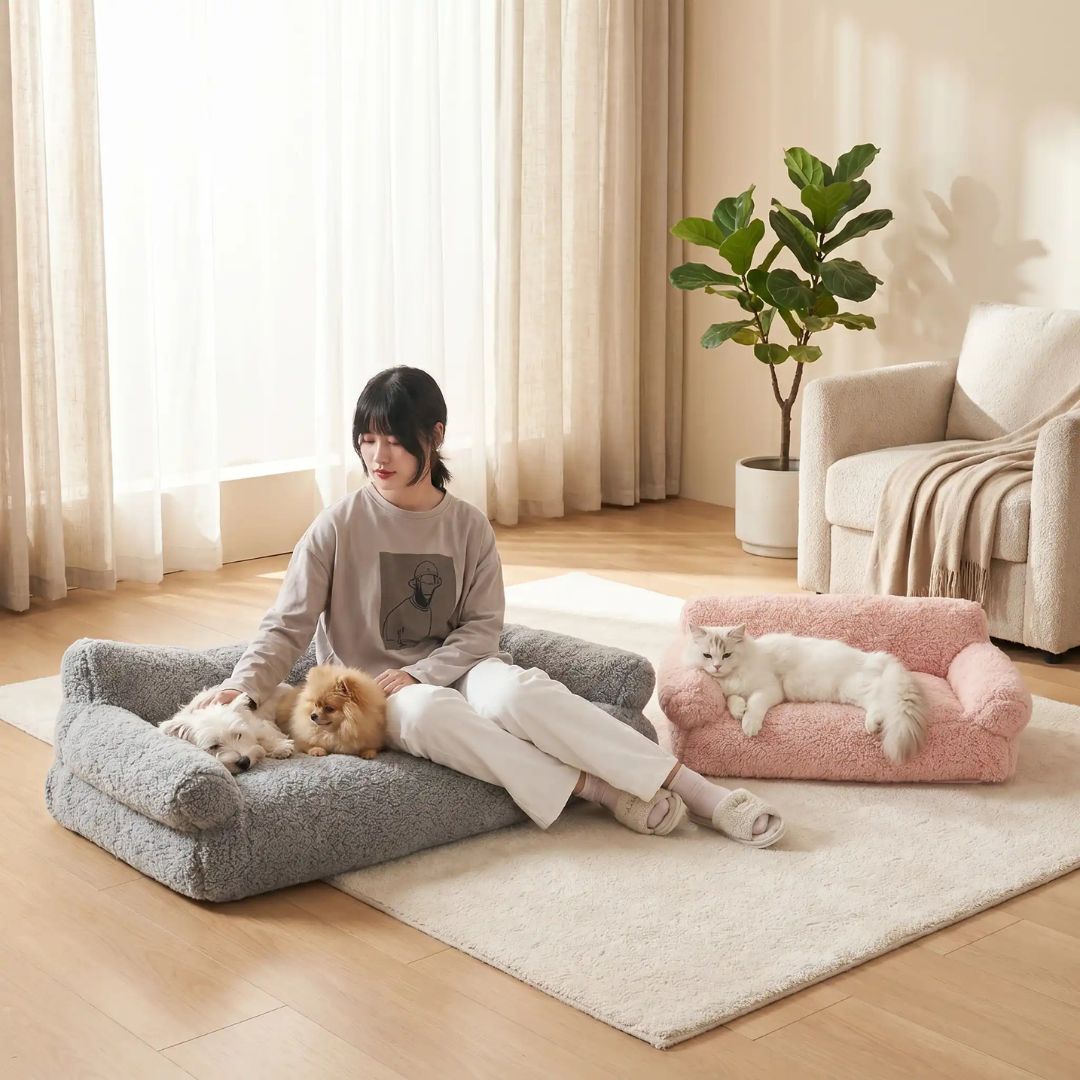 Canapés-pour-chiens-xxl-120-cm, déhoussable, antidérapant et lavable en machine, en peluche éponge douce et rembourrage 100% coton haute densité, housse 100% imperméable, avec 2 dossiers et 2 accoudoirs pour chien de 0 à +40 kg- une maitresse assise avec ses petits chiens sur le canapé xxl pour chien et chiot de grande race de couleur gris avec son chat allongé confortablement sur le canapé rose dans le salon de l'appartement
