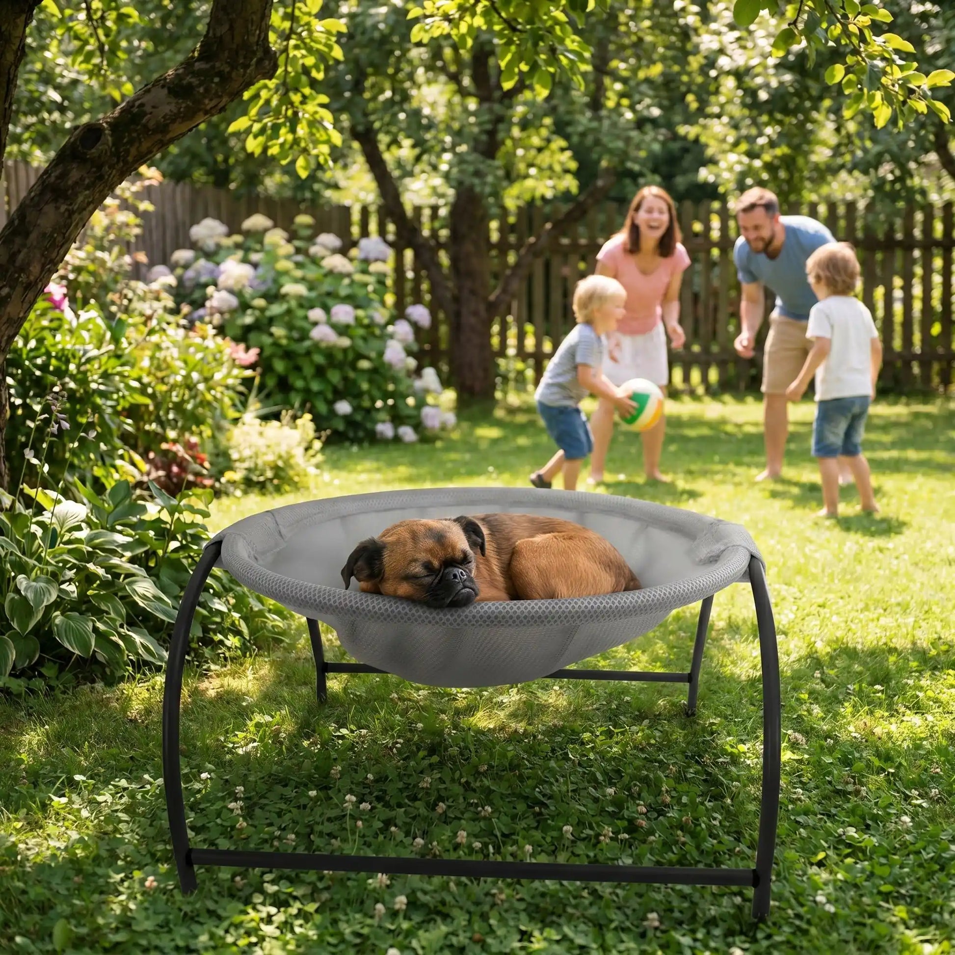 Hamac-pour-chien-chiot-exterieur et interieur en maille respirante et hypoallergénique, structure solide en acier avec 3 pieds et renfort par de barres latérales, cerceau central entourant et apaisant pour les chiot et chien de petite taille et de races naines de 0 à 3 kilos max , lit surélevé et idéal pour l'été-un chien de race miniature qui dort paisiblement niché dans le hamac rond avec pied surélevé sous l'arbre du jardin avec sa famille qui profite de l'été dehors