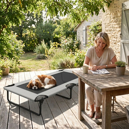 Hamac pour chien de 107 cm, en tissu oxford 100% imperméable et maille respirante et structure en acier robuste, polyvalent pour une utilisation interieur et exterieur, surélevé avec 3 barres transversales et patin anti rayure pour une stabilité pour chiot et chien de petite à moyenne taille jusqu'à 30 kilos-un chien à poils long et épais qui dort couché au frais sur son lit surélevé gris foncé au pied de sa maitresse qui joue aux mots croisée sur la table en bois de la terrasse de la maison dans le sud 