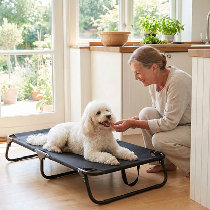 Lit-pliant-chien-camp-4-tailles du S au XXL ppur chiot et chien de toute races , imperméable en tissu teslin ultra résistant et respirant , rafraichissant en été, surélevé avec ses pieds et sa structure en métal robute avec barre au milieu pour une maille qui ne s'affaisse pas, avec poignée de transport, léger et compact pour un lit interieur et extérieur- un caniche confortablement couché sur son lit surélevé hamac noir de taille M et sa maitressequi lui donne une friandise dans la cuisine en été 
