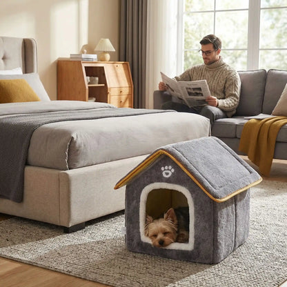 Maison-chien-intérieure-chiot-mini, pliable, légère et transportable, avec matériau isolant efficace contre le bruit et du froid, niche antidérapante, lavable en machine avec toit amovible, de couleur grise pour chiot et chien de 0 à 5 kg, disponible en taille XS et S- un petit chien qui dort paisiblement dans sa petite maison grise avec le design de pattes de chien mignon sur l'entrée de la niche avec toit rigide dans la location de vacance avec son maitre qui lit le journal sur le canapé 