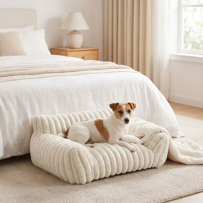 Mini-canapé-chien-chiot-déhoussable, lavable en machine ,coton, housse en peluche douce au toucher velours, rembourrage ergonomique en mousse à mémoire de forme pour un soutien optimal, disponible en  couleurs blanc et rose et 3 tailles S, M et L pour chiot et chien de petite taille de 0 à 10 iols max-un chien de petite race couché confortablement avec les deux accoudoirs et dossier rembourrés entourant du mini fauteuil pour chien de couleur blanc crème au pied du lit de sa maitresse dans la chambre 
