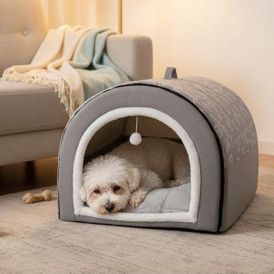 Niche-pour-chiot-anti-stress, en forme de grotte ou d'igloo semi-fermé pour une bonne isolation sonore qui protège du froid, un refuge sécurisé avec ubn coussin intérieur amovible et lavable en machine, structure robuste et pliable 100% imperméable et résistant, antidérapant, transportable avec sa poignée intégrée pour chiot de 0 à 15 kilos-un chiot coton de tuléar qui dort dans la niche chaude et semi fermée de couleur grise dans le salon de l'appartement