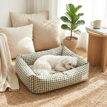 Panier chien et chiot rectangulaire avec rebord 360° surélevé en tissu pour chien de petite taille, 100% coton haute densité avec coussin intérieur amovible double-face été/hiver lavable en machine avec fond imperméable et antidérapant, design à carreaux vichy chic et frais pour un lit 4 saisons pour chien et chiot de 0 à 9 kilos-un Bichon maltais qui dort paisiblement niché contre les bords du panier rectangulaire rembourré et confortable à carreaux vichy de couleur vert sur le sol de la chambre 