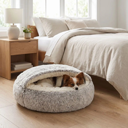 Panier-couvert-chien-chiot douillet, lit en peluche rond avec toit souple qui sert de couverture cousue au panier rond et profond comme un donut au rembourrage 100% coton haute densité entourant et ergonomique pour un effet apaisant et anti-stress, chaud pour les chiots et chiens de petite taille de 0 à 5 kg, anxieux ou frileux-un chien de petite taille qui dort niché dans son panier rond gris avec le toit reposant sur sa tête pour un effet calmant qui protège du froid au pied du lit de sa maitresse  