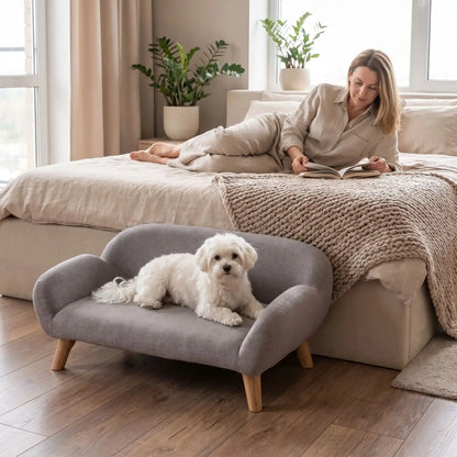 Petit-canape-pour-chien-chiot-design de couleur gris, dossier et accoudoirs arrondis dans un style scandinave, tissu résistant aux griffes, rembourrage 100% coton haute densité pour un confort optimal, structure en bois massif et surélevé avec 4 pieds en bois massif pour chiot et chien de 0 à 8 kilos-un petit chien couché confortablement sur son canapé gris design sur pied en bois massif et rembourrage en éponge coton ergonomique au pied du lit de sa maitresse alllongée qui lit dans la chambre 