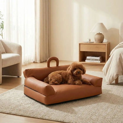 Petit-canapé-pour-chien-chiot-imperméable, déhoussable lavable en machine, en Cuir végan et rembourrage 100% coton haute densité et résilience, avec accoudoirs, dossier et assise ergonomique, avec poignée de transport, antidérapant et anti humidité, durable solide et résistant aux griffes pour chiot, petit chien et race naine de 0 à 10 kilos- un caniche nain couché sur  son mini canapé marron dans la chambre de la maison