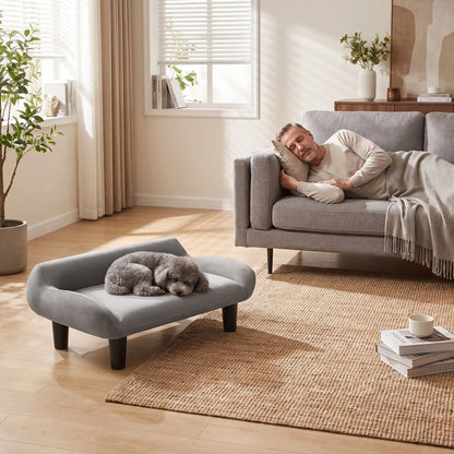 Petit-fauteuil-pour-chien-chiot-design de couleur gris, dossier et accoudoirs arrondis dans un style scandinave, velours résistant aux griffes,rembourrage 100% coton haute densité pour un confort optimal,structure en bois massif et surélevé avec 4 pieds en bois massif pour chiot et chien de 0 à 8 kilos-un chien de race naine qui dort paisiblement sur son canapé gris design sur pied en bois massif et rembourrage en éponge coton ergonomique au pied de son maitre qui fait la sieste sur son propre canapé 