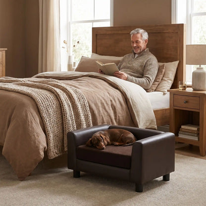 Petit-fauteuil-pour-chien-chiot-design en bois massif robuste  et cuir vegan sur 4 pieds avec coussin interieur épais rembourré 100% coton haute densité et ergonomique amovible déhoussable et lavable en machine, pied antidérapant et anti rayure pour chiot et chien de petite taille de 0 à 5 kilos-un chien de race miniature qui dort paisiblement sur le mini canapé design de couleur marron à côté du lit de son maitre qui lit son mivre allongé dans la chambre lifestyle de l'appartement