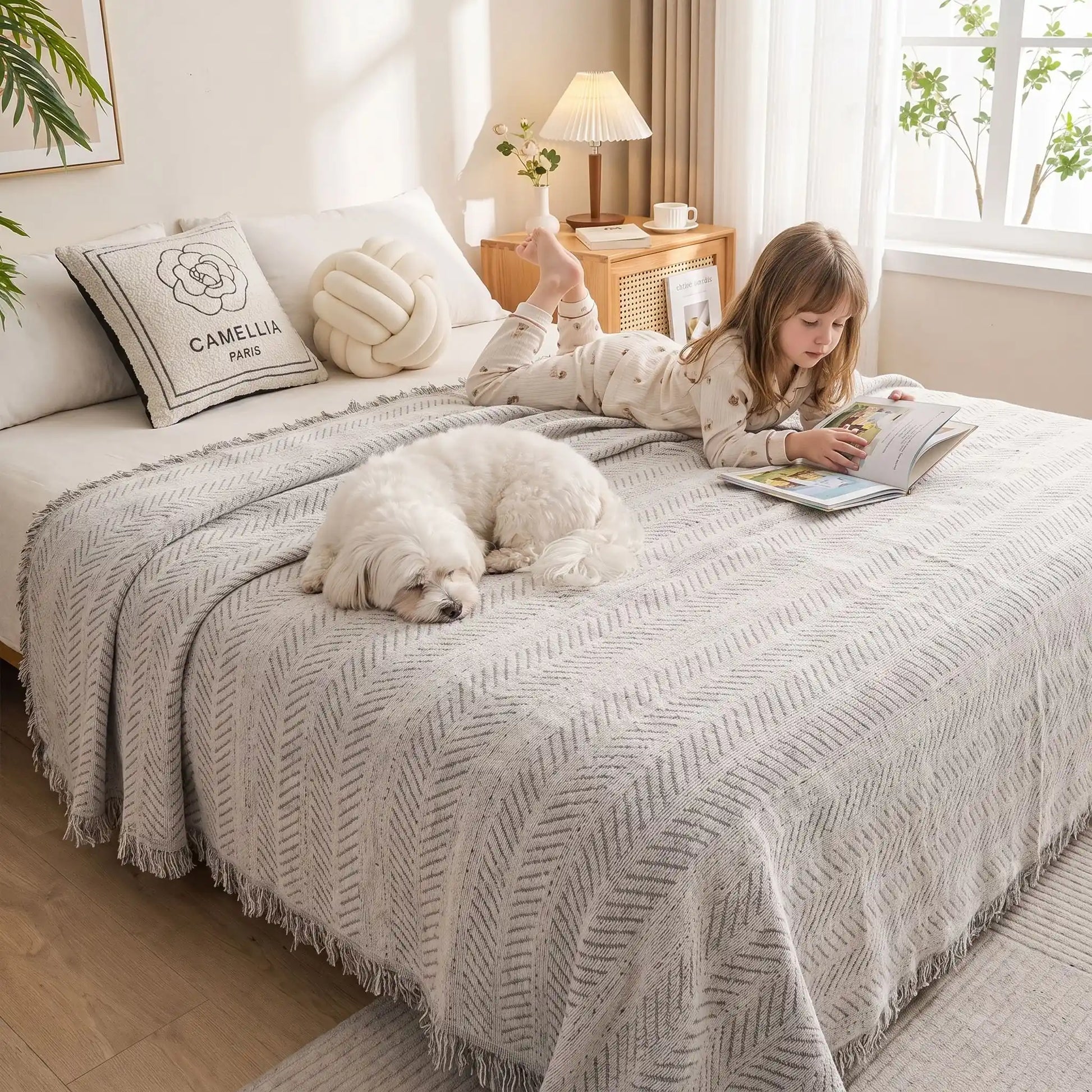 Protection-canape-chien-anti-poils-résistante aux griffes, tissage chenille coton et polyester, écoresponsable avec franges pour un style bohème chic de couleur gris clair, dispoinble en 4 tailles pour fauteuil/canapé une place, deux places, 3 places et 4 places, lavable en machine, doux respirant confortable et 4 saisons- un chien de petite taille qui dort à côté d'une petite fille qui lit son livre allongés tout deux sur le plaid de protection sur le lit deux places des maitres dans la chambre 