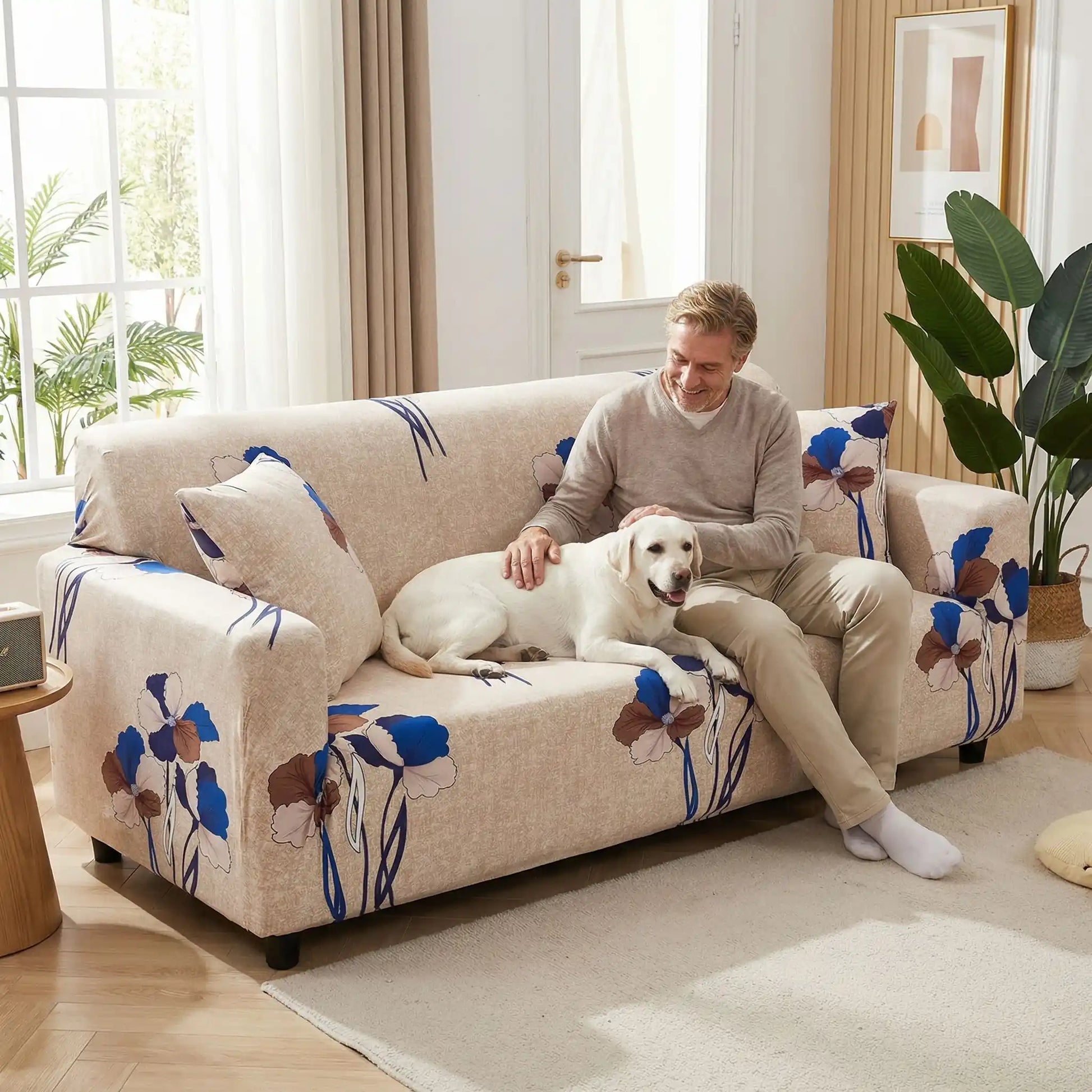 Protection-canape-chien-anti-poils-anti accident, lavable en machine, haute élasticité et resistante aux griffes et à l'usure, universelle en 4 tailles pour fauteuil, canapé de 2/3/4 places, avec élastique et bloque plis- design floral délicat- un maitre assis confortablement sur le canapé qui caressse tendrement son grand chien couché à côté de lui sur le canapé dans le salon de la maison